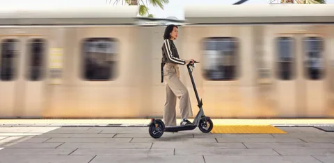Femme sur une Niu KQi Air sur un quai de gare, train flou derrière